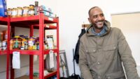 A photo of Voices in Exile service user, Mohammed, volunteering at the food bank. A man stands in front of a shelf full of food smiling